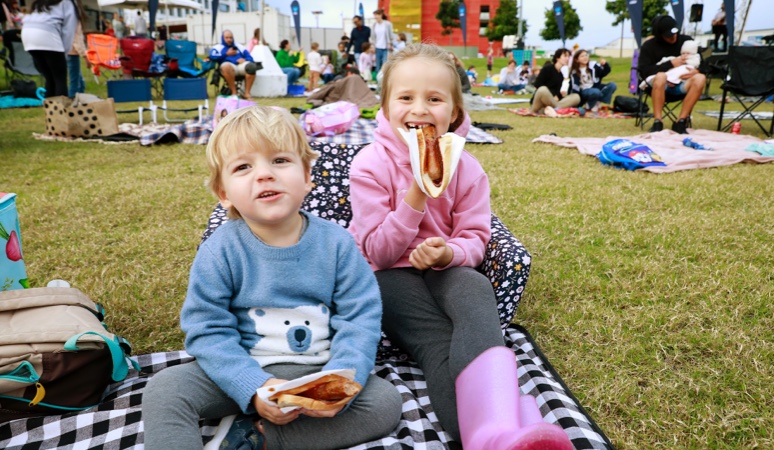 Two children on a picnic rug eating sausage in bread and groups of people sitting down in the background.